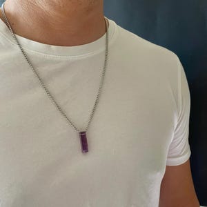 May include: A silver-toned metal chain necklace featuring a rectangular, deep purple amethyst pendant. The pendant is vertically oriented and suspended from a simple link chain that rests against a plain white t-shirt, creating a clean look.