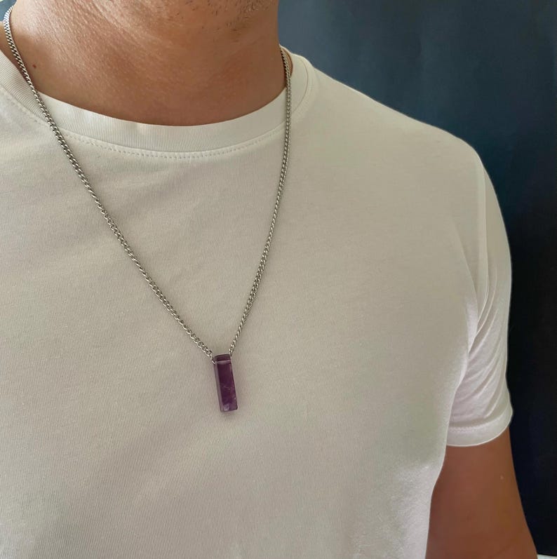 May include: A silver-toned metal chain necklace featuring a rectangular, deep purple amethyst pendant. The pendant is vertically oriented and suspended from a simple link chain that rests against a plain white t-shirt, creating a clean look.