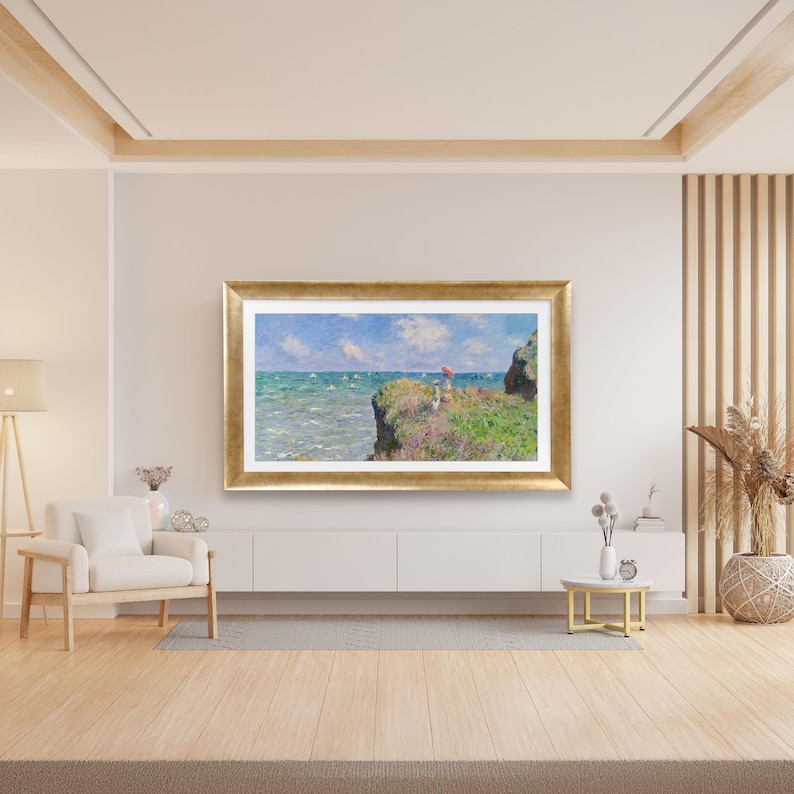 Puede incluir: Una pintura de paisaje enmarcada con un marco dorado representa una escena costera. La obra de arte muestra un acantilado con vistas a un mar turquesa bajo un cielo azul con nubes blancas. Figuras con un paraguas rojo est&aacute;n en el acantilado.