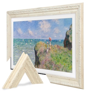May include: A framed print of a coastal landscape painting with a white ornate frame. The artwork depicts a cliffside scene with figures, the ocean, and a blue sky. A detached corner of the frame is visible.