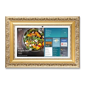 Puede incluir: Una pantalla digital rectangular con un marco dorado y ornamentado. La pantalla muestra una receta de ensalada y el pronóstico del tiempo. La receta es para ensalada de feta y rúcula. El marco tiene tallas detalladas.