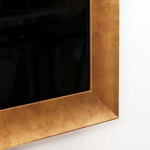 Puede incluir: Un espejo rectangular con un marco ancho y texturizado de color dorado. El marco tiene una apariencia ligeramente desgastada. La superficie del espejo es negra, reflejando el entorno. El marco est&aacute; montado en una pared blanca.