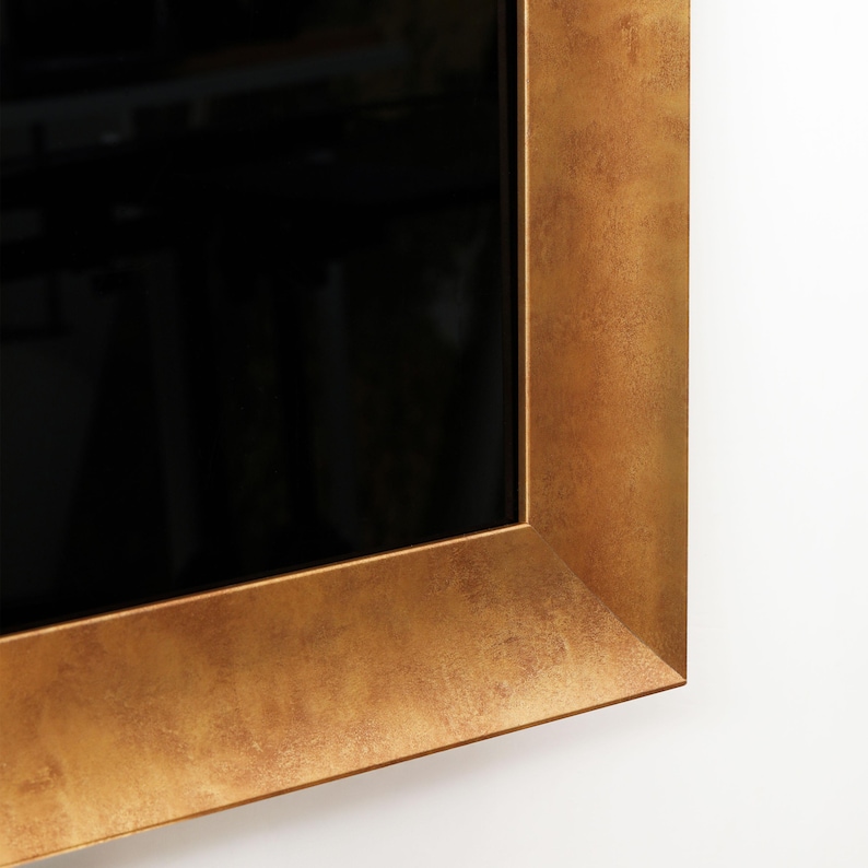Puede incluir: Un espejo rectangular con un marco ancho y texturizado de color dorado. El marco tiene una apariencia ligeramente desgastada. La superficie del espejo es negra, reflejando el entorno. El marco est&aacute; montado en una pared blanca.