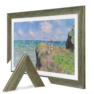 May include: A framed print of a coastal landscape painting. The artwork depicts a cliffside scene with figures, sailboats, and a blue sky. The frame is a distressed green with a rustic appearance, and a detached corner is visible.
