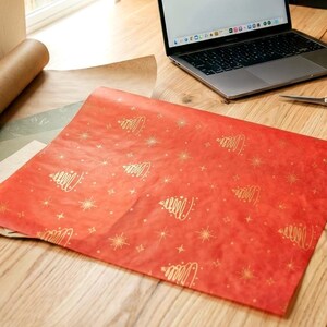 Peut inclure: Un ordinateur portable est posé sur un bureau en bois, à côté d'un papier cadeau rouge orné d'un motif de sapins dorés. Un rouleau de papier brun et des ciseaux sont également présents. La scène est éclairée par la lumière naturelle d'une fenêtre.
