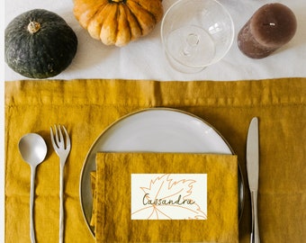 Puede incluir: Una tarjeta de lugar blanca con un contorno marrón de una hoja y el nombre "Cassandra" escrito en cursiva marrón. La tarjeta de lugar está sobre una servilleta de lino amarilla, que está sobre un plato blanco. Hay tenedores y cucharas de plata en la mesa.