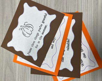 May include: A stack of gratitude cards with a pumpkin illustration. The cards have a brown border with white, scalloped-edge inserts. Text on the cards includes prompts like "Name three things you're grateful for" and "What that brings".