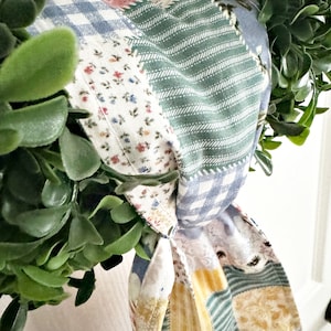May include: A patchwork fabric wreath with a green, blue, and yellow color scheme. The fabric is tied into a bow and the wreath is decorated with green leaves.