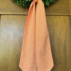 May include: A long, fabric ribbon featuring an orange and white gingham check pattern. The ribbon is tied in a simple knot at the top, with two long, straight ends hanging down vertically, displaying a consistent checkered textile design.