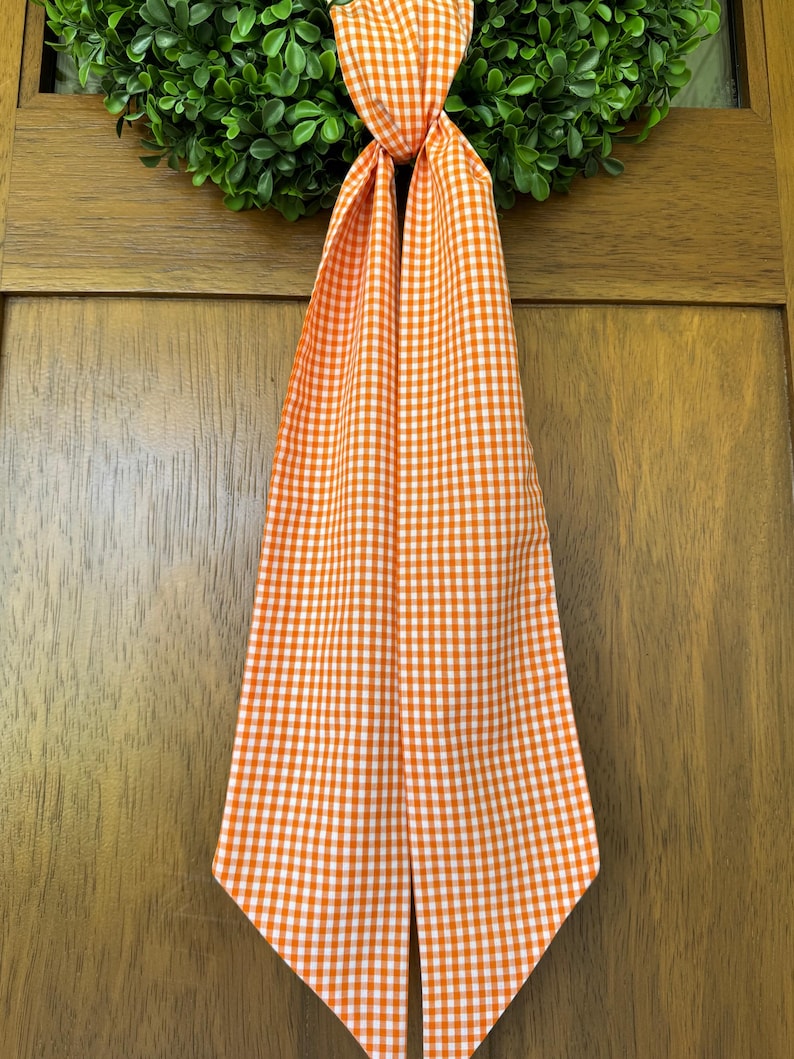 May include: A long, fabric ribbon featuring an orange and white gingham check pattern. The ribbon is tied in a simple knot at the top, with two long, straight ends hanging down vertically, displaying a consistent checkered textile design.