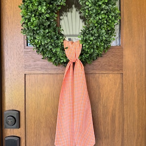 May include: A circular wreath composed of dense, vibrant green faux boxwood leaves. The wreath is adorned at the bottom with a long, hanging fabric ribbon featuring a classic orange and white gingham check pattern that drapes downward.