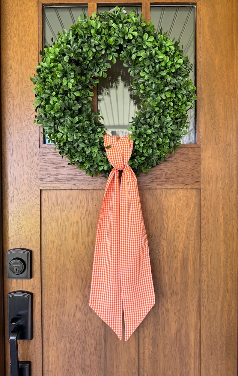 May include: A circular wreath composed of dense, vibrant green faux boxwood leaves. The wreath is adorned at the bottom with a long, hanging fabric ribbon featuring a classic orange and white gingham check pattern that drapes downward.