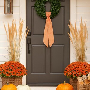 May include: A dark gray front door features a green wreath with an orange gingham ribbon. The entryway includes two woven baskets filled with orange chrysanthemums and tall dried grasses, alongside several orange and white pumpkins and gourds.