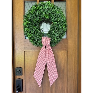 May include: A green boxwood wreath with a red and white gingham ribbon hanging on a wooden door. The wreath is round and full, with small, dense leaves. The ribbon is tied in a bow and hangs down.