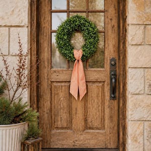 May include: A circular green boxwood wreath hangs on a rustic wooden door with a glass pane. The wreath is adorned with a long, orange and white gingham ribbon tied in a bow at the bottom, with two long tails hanging down the center of the door.