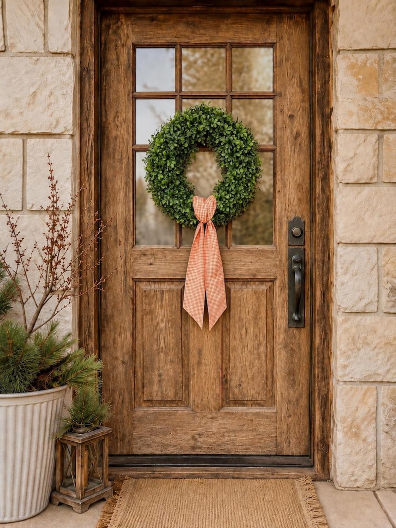 May include: A circular green boxwood wreath hangs on a rustic wooden door with a glass pane. The wreath is adorned with a long, orange and white gingham ribbon tied in a bow at the bottom, with two long tails hanging down the center of the door.