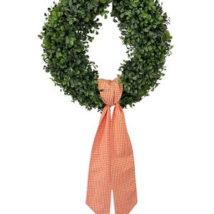May include: A circular wreath constructed from dense, dark green artificial boxwood foliage. The wreath is adorned at the bottom with a long, hanging fabric ribbon featuring a small-scale orange and white gingham checkered pattern.