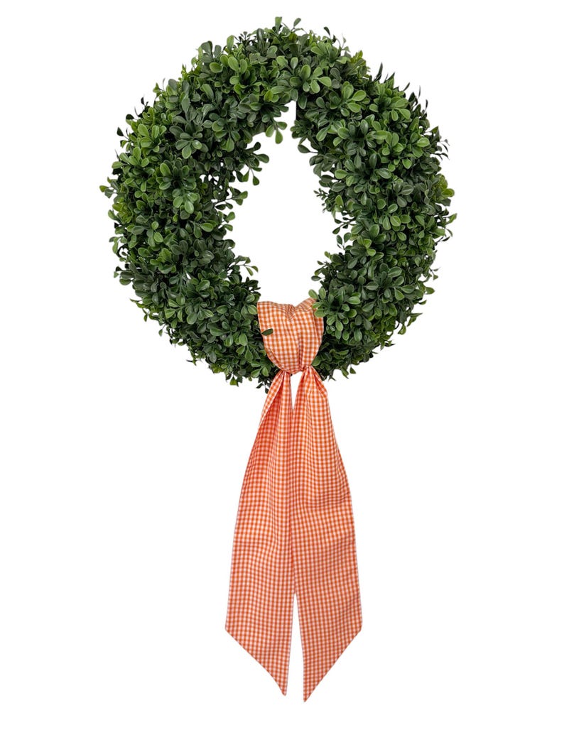 May include: A circular wreath constructed from dense, dark green artificial boxwood foliage. The wreath is adorned at the bottom with a long, hanging fabric ribbon featuring a small-scale orange and white gingham checkered pattern.