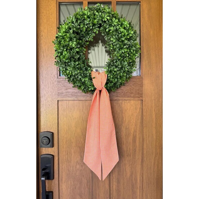 May include: A circular wreath composed of dense, vibrant green boxwood leaves. Attached to the bottom center of the wreath is a long, flowing fabric ribbon featuring a classic orange and white gingham checkered pattern that hangs vertically.