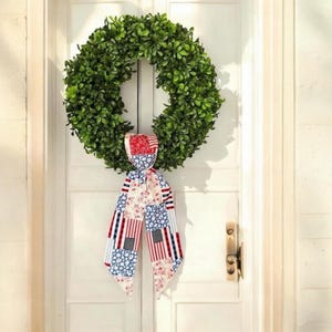 May include: A green, leafy wreath hangs on a white door. A decorative ribbon with a patchwork design of red, white, and blue stars and stripes is tied to the wreath. The door has a gold-colored handle.
