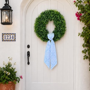 May include: A circular green boxwood wreath hangs on a white arched door. The wreath is adorned with a long, light blue ribbon featuring a white floral pattern that hangs down from the center. The door has a black handle and a black lock mechanism.