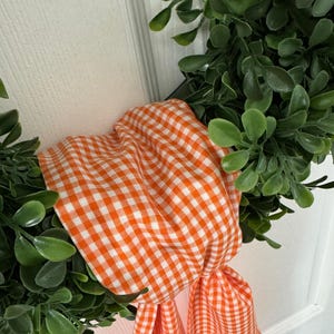 May include: A decorative wreath featuring dense, rounded green leaves arranged in a circular shape. An orange and white gingham fabric ribbon is tied around the foliage, with two fabric ends hanging down from the center of the wreath.
