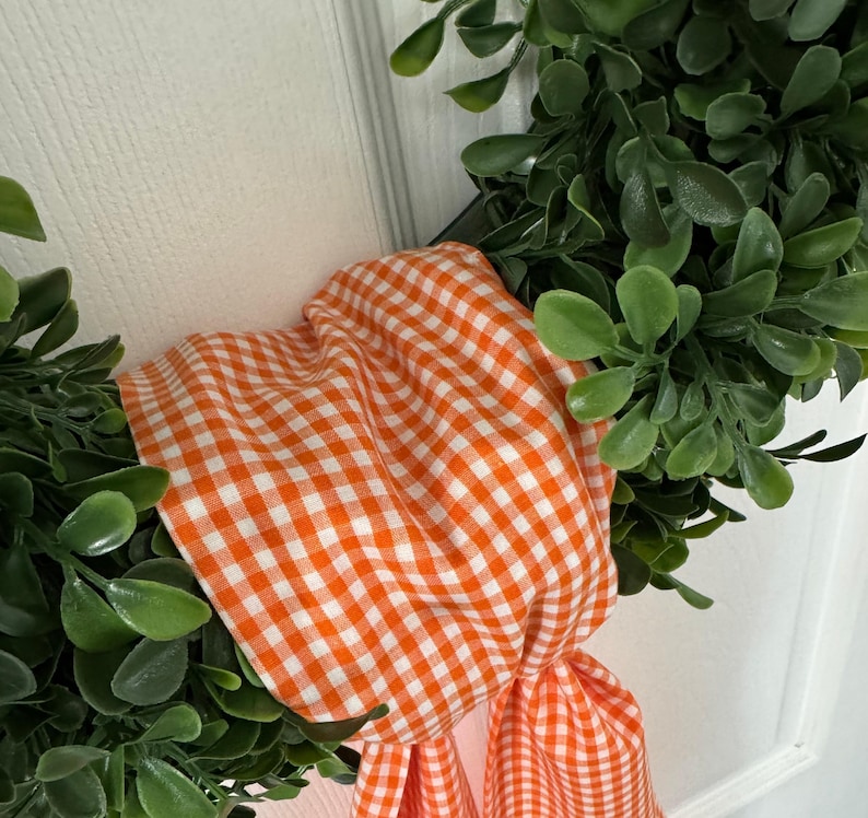 May include: A decorative wreath featuring dense, rounded green leaves arranged in a circular shape. An orange and white gingham fabric ribbon is tied around the foliage, with two fabric ends hanging down from the center of the wreath.