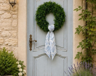 May include: A light blue wooden door with a dark metal handle and a green boxwood wreath hanging in the center. The wreath is adorned with a long, hanging fabric ribbon featuring a blue and white floral toile pattern, centered on the door.