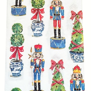 May include: White paper napkin with a repeating pattern of nutcrackers, topiary trees, and decorative pots. The nutcrackers are in blue and red uniforms, standing on blue drums. The topiary trees are green with red bows, and the pots are blue and white.