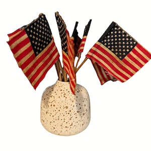 May include: A white ceramic vase with brown speckles holds a bouquet of small American flags. The flags are made of red, white, and blue fabric with a distressed look.