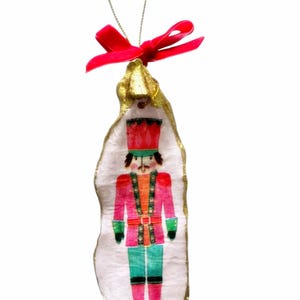 May include: A pink and gold Christmas ornament shaped like a nutcracker. The nutcracker is wearing a red and green uniform with a gold belt and a black hat. The ornament has a red ribbon attached to the top.
