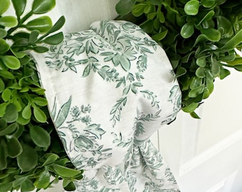 May include: A white fabric ribbon with a green floral pattern is tied around a green leafy wreath. The wreath is hanging on a white door.