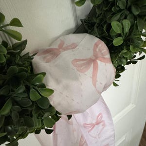 May include: A pink fabric wreath with a white background and pink bows printed on it.