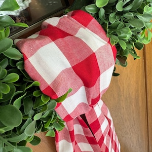 May include: Red and white checkered fabric bow tied around a green wreath. The bow is made of a soft, woven fabric with a classic gingham pattern.