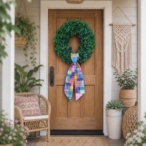May include: A wooden front door with a green wreath and a colorful patchwork ribbon. A woven chair with a patterned pillow sits to the left, and potted plants and a macrame wall hanging decorate the porch. A natural fiber rug is on the floor.