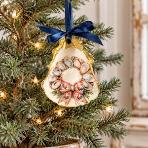 May include: A decorative oyster shell ornament featuring a painted wreath of smaller shells and a central bow with rooster motifs. The shell edges are gilded in gold, and it hangs from a dark blue satin ribbon tied in a bow at the top.