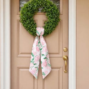 May include: A green boxwood wreath hangs on a tan door, adorned with a white ribbon printed with pink and green floral patterns. The door has a brass handle and a small window at the top, creating a welcoming entrance.