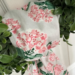 May include: A white fabric wreath with a floral pattern featuring pink and green hydrangeas. The wreath is decorated with green leafy branches.