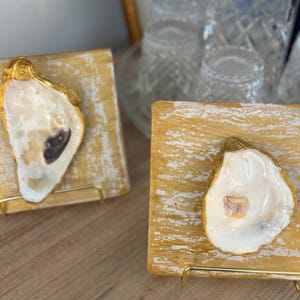 May include: Two decorative oyster shell art pieces. Each shell is mounted on a square, distressed, gold-painted wooden block. The oyster shells are white with gold trim and sit on a gold metal stand.