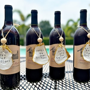 May include: Four bottles of red wine with decorative oyster shell tags. Each tag has the phrase "corks are for quitters" written on it. The tags are gold-edged and attached with twine and wooden beads.