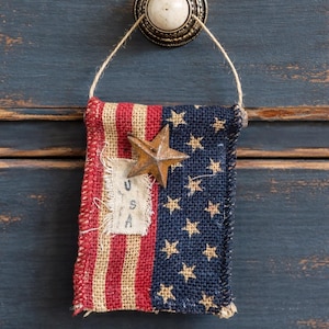 May include: A rustic burlap flag with red and white stripes and a blue field with gold stars. A metal star is affixed to the flag. The flag is hanging from a drawer pull, evoking a vintage, patriotic theme.