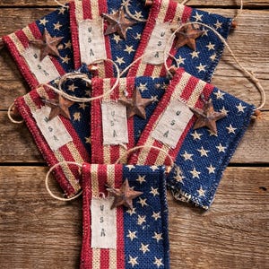 May include: A collection of patriotic burlap banners with a rustic aesthetic. Each banner features a design reminiscent of the American flag, with red and white stripes, blue star-studded fields, and a metal star accent. The word "USA" is printed on a central strip.