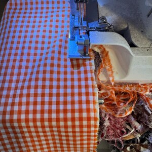 May include: A white sewing machine serger is actively stitching the edge of a piece of orange and white gingham fabric. The machine needle and presser foot are visible, with frayed fabric scraps accumulating in a pile near the machine base.