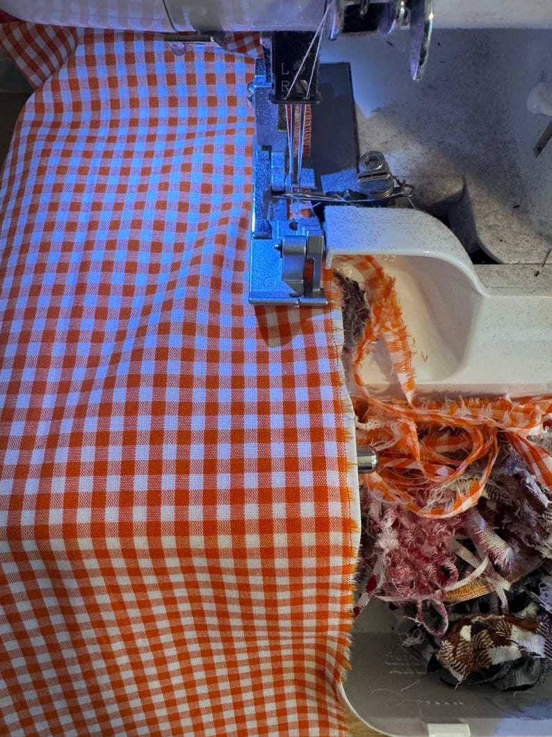 May include: A white sewing machine serger is actively stitching the edge of a piece of orange and white gingham fabric. The machine needle and presser foot are visible, with frayed fabric scraps accumulating in a pile near the machine base.