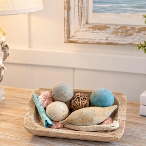 May include: A rectangular wooden dough bowl filled with decorative items including two smooth spheres in grey and blue, a small woven rattan ball, a light blue piece of coral, a tan seed pod, and several small seashells in shades of pink and white.