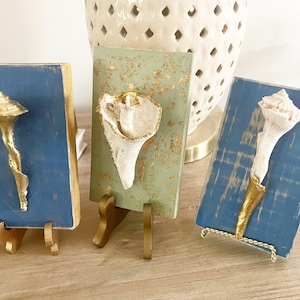 May include: Three small wooden plaques with gold accents, each featuring a different seashell. The plaques are displayed on gold metal stands.