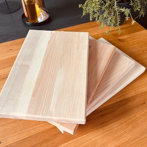 Puede incluir: Tres tablas de cortar de madera de diferentes tamaños, apiladas una encima de la otra. Las tablas son de madera clara con un patrón de veta natural.