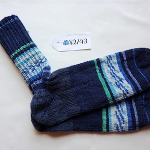 May include: A pair of hand-knitted navy blue socks featuring horizontal stripes in shades of light blue, white, and bright green. A white rectangular tag is attached, displaying the text 42/43 with a small blue icon of two socks.