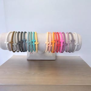 May include: A display of colorful silicone bracelets with a cross charm. The bracelets are arranged in a rainbow pattern on a white stand.