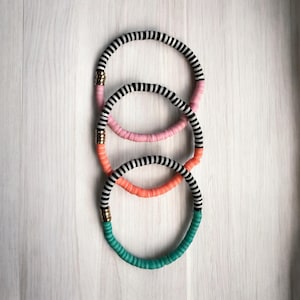 Op de afbeelding: Drie kleurrijke kettingen met een zwart-wit gestreept ontwerp. De kettingen zijn verticaal gestapeld, met roze, oranje en turquoise accenten. Goudkleurige metalen sluitingen verbinden de gekleurde secties.
